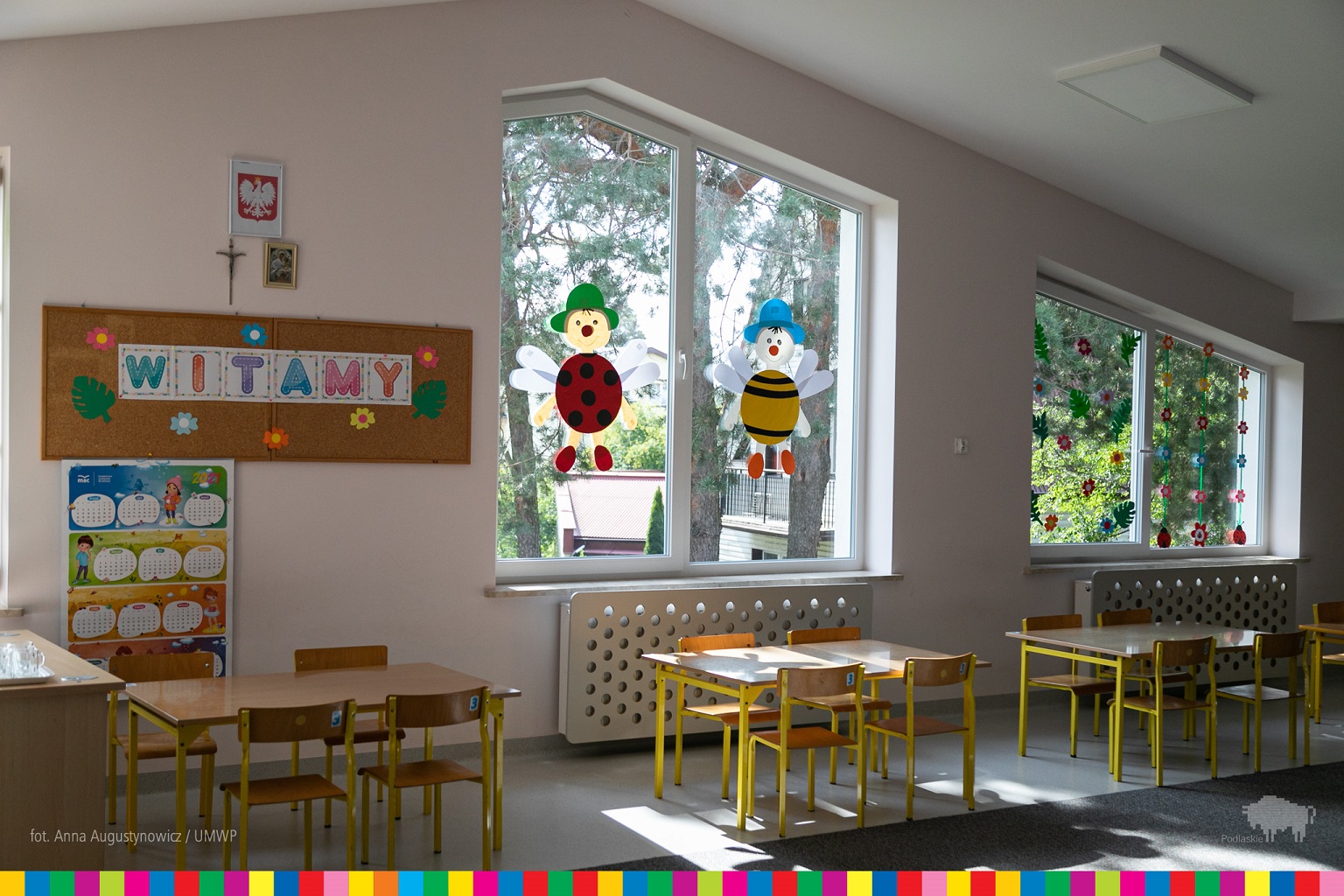 Wnętrze sali w przedszkolu. W oknach kolorowe naklejki, dziecięce stoły i krzesła.