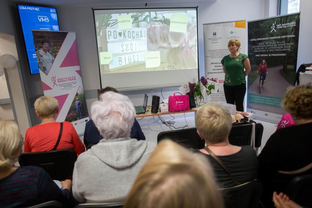 Przedstawicielka  Białostockiego Centrum Onkologicznego stoi przed grupą słuchaczy i prezentuje informacje o profilaktyce raka piersi