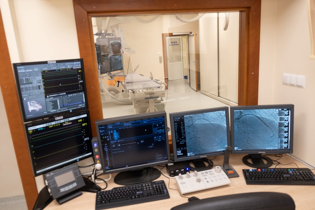 Kabina z której wykonuje się badanie angiografem, na biurku stoi pięć ekranów komputerowych, za szybą angiograf